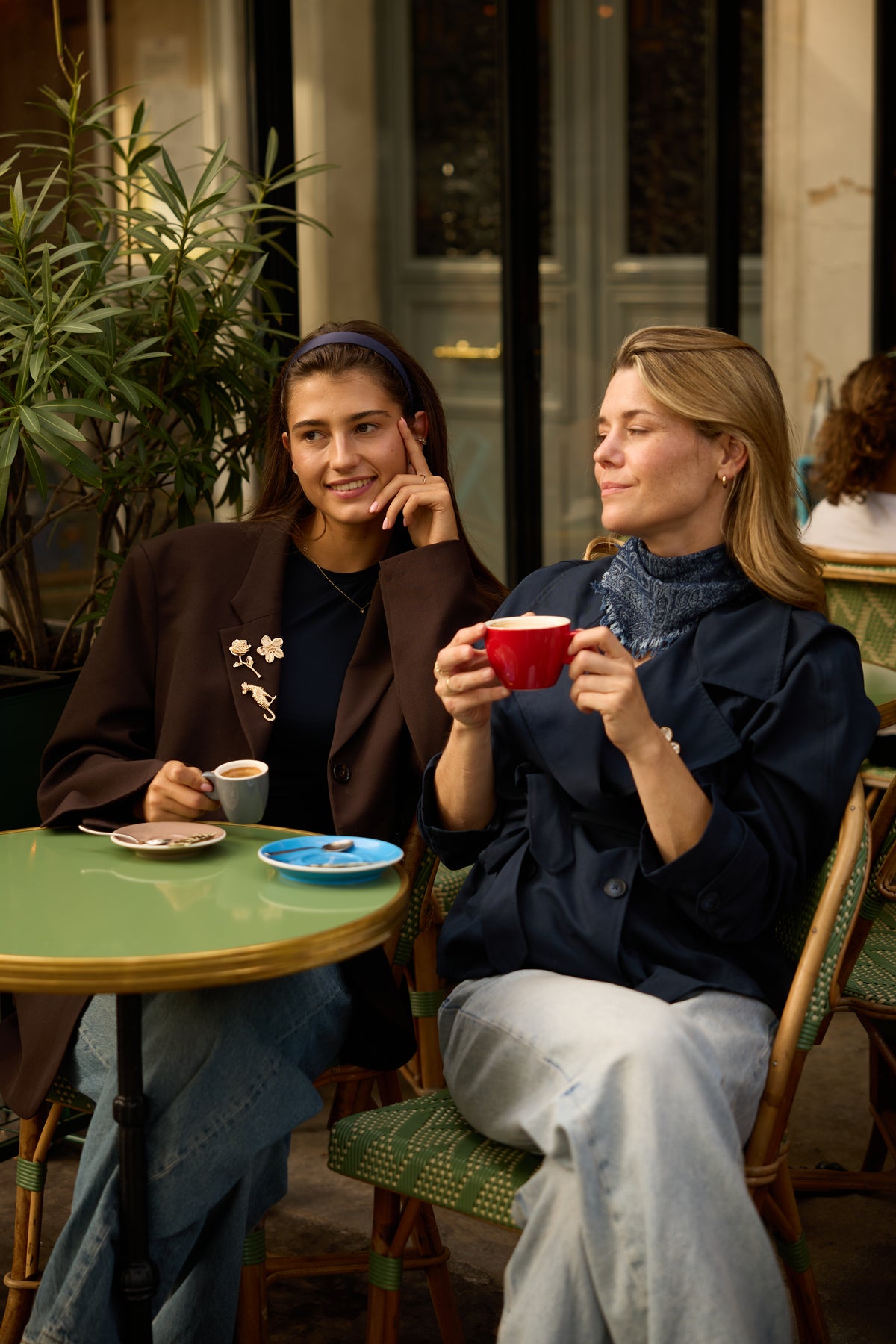 Lara Flower gold magnetic brooch on the left model's brown blazer alongside Juliette rose and Santos leopard brooches at a Paris cafe; each design is a separate product.