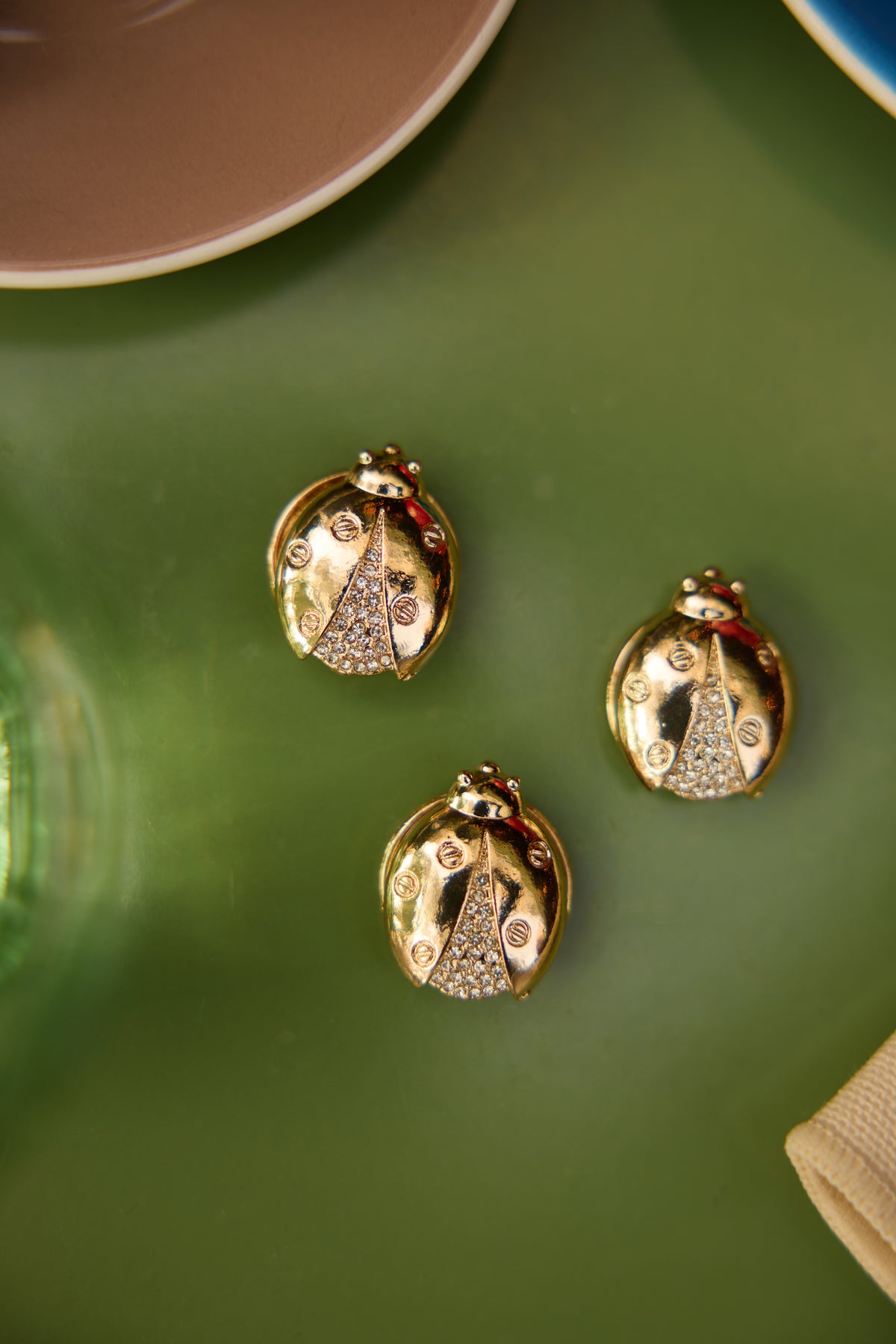 Close styling image of three separate Olivia gold ladybird magnetic brooches on a green cafe table; this product is sold individually.