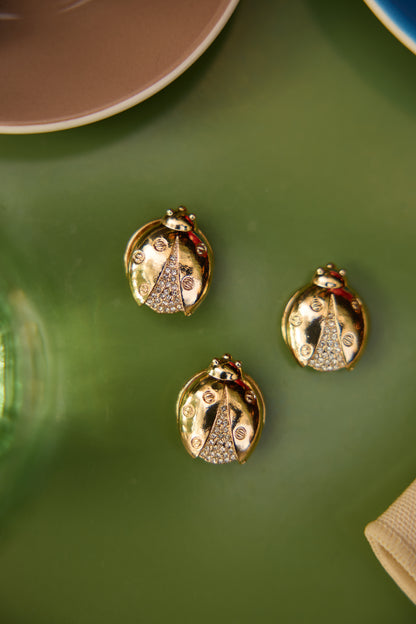 Close styling image of three separate Olivia gold ladybird magnetic brooches on a green cafe table; this product is sold individually.